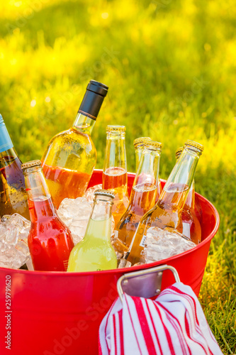 Cold beverages wine, beer and soda on ice in red bucket pail tub on green grass. Summer outdoor backyard garden party picnic.