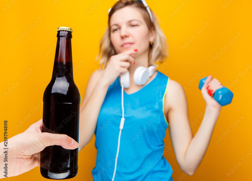 Sportive girl in headphones with dumbbell in handin front of hand with bottle of beer. Sportive girl in headphones with dumbbell in handin front of hand with bottle of beer.