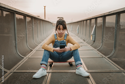 Stylish teen in jean overall sitting on skateboard and using mobile phone on footpath in city