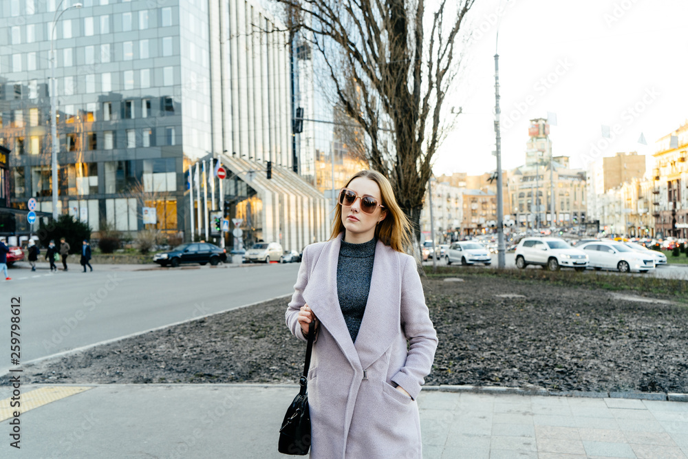 Beautiful young caucasian woman walking along the street in the city. Stylish female model wearing sunglasses walking on city street.