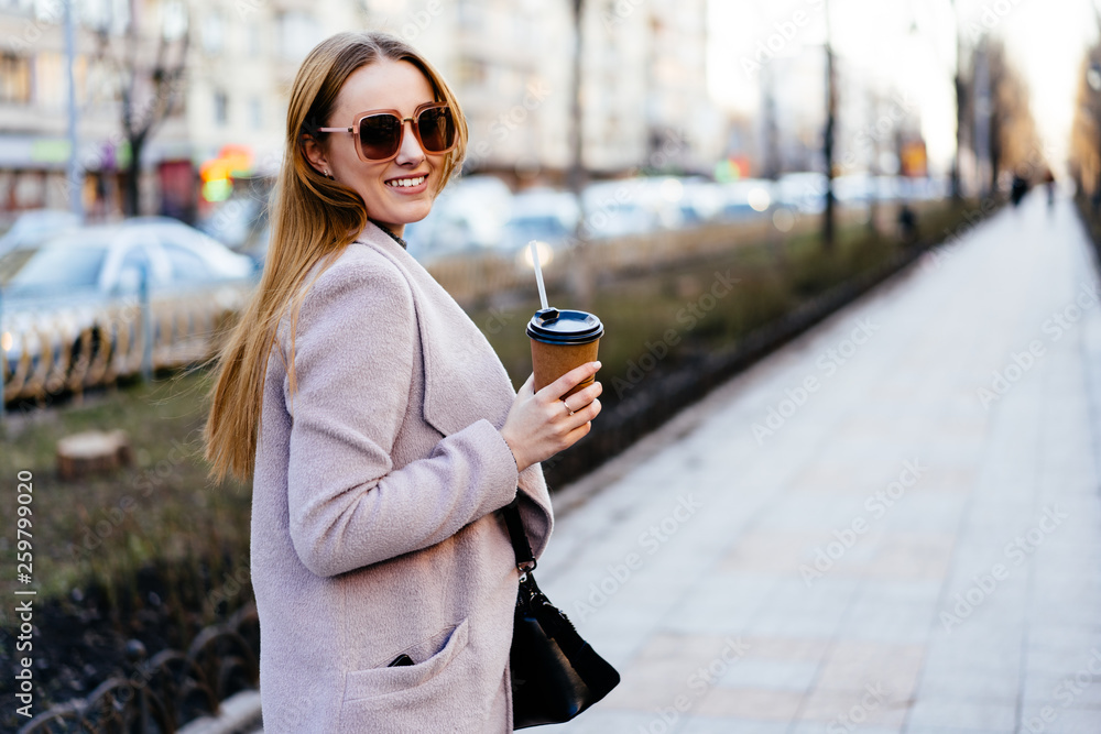 Beautiful young caucasian woman walking along the street in the city. Stylish female model wearing sunglasses with disposable cup walking on city street. Beautiful young caucasian woman walking along the street in the city. Stylish female model wearing sunglasses with disposable cup walking on city street.