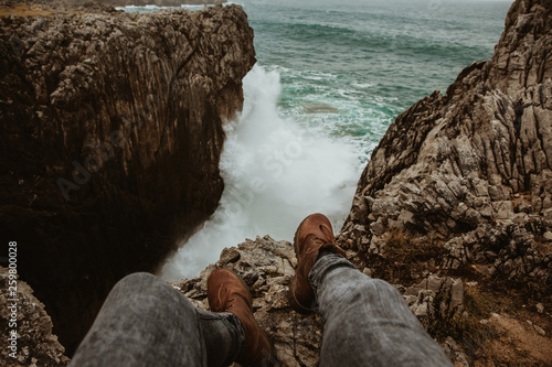 Crop legs of human sitting on top of stone near stormy sea in Bufones de Pria, Asturias, Spain photo