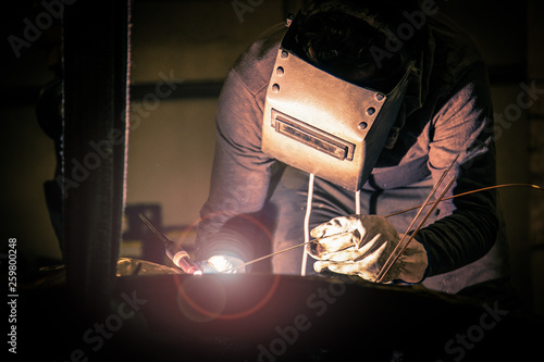 Side view of male in welding helmet working with metal product in obscurity on little foundry