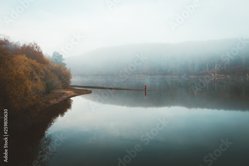 Picturesque landscape of beautiful silent lake with reflection of forest and lonely tourist in thick fog photo