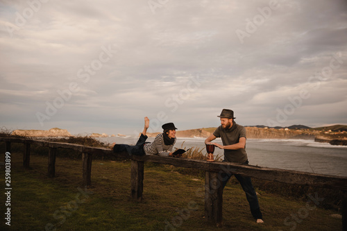 Young guy in hat playing on hand drum near elegant lady in cap with notepad and pencil lying on seat near coast of sea and cloudy sky