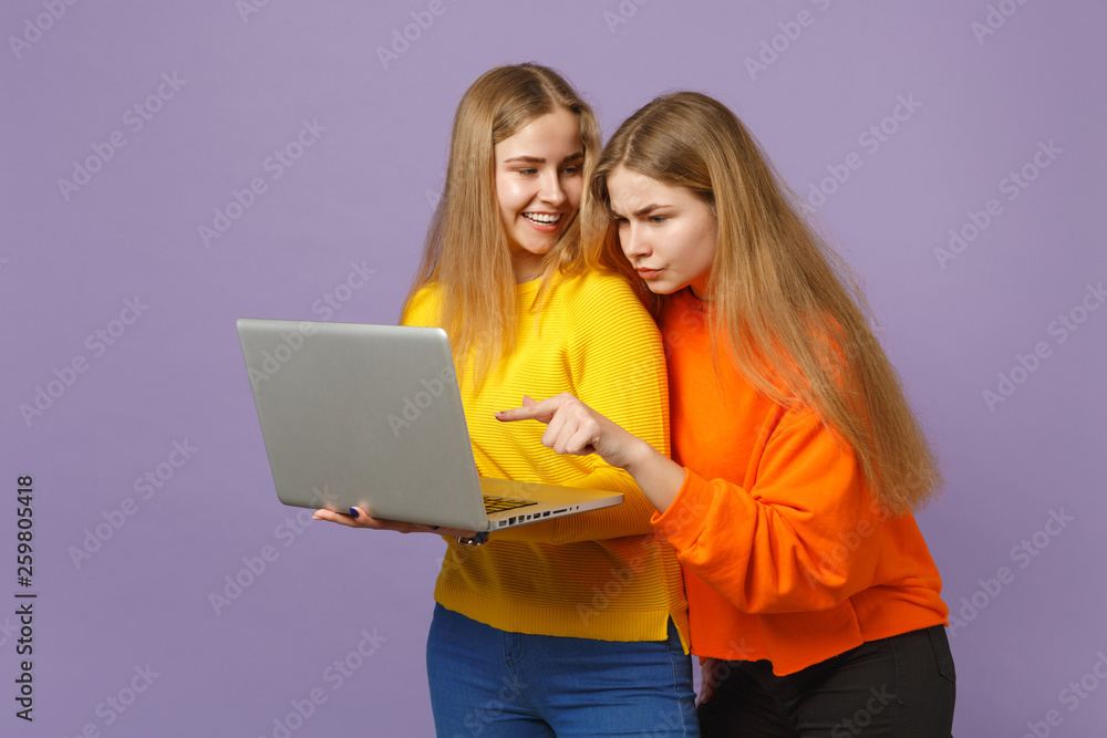 Two funny young blonde twins sisters girls in vivid colorful clothes holding, using laptop pc computer isolated on pastel violet blue background. People family lifestyle concept. Mock up copy space.