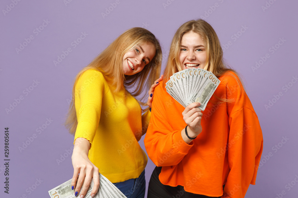 Two happy blonde twins sisters girls in colorful clothes holding fan of money in dollar banknotes, cash money isolated on violet blue background. People family lifestyle concept. Mock up copy space. Two happy blonde twins sisters girls in colorful clothes holding fan of money in dollar banknotes, cash money isolated on violet blue background. People family lifestyle concept. Mock up copy space.