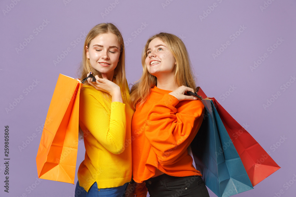 Two pensive young blonde twins sisters girls in vivid clothes holding package bag with purchases after shopping isolated on violet blue background. People family lifestyle concept. Mock up copy space. Two pensive young blonde twins sisters girls in vivid clothes holding package bag with purchases after shopping isolated on violet blue background. People family lifestyle concept. Mock up copy space.