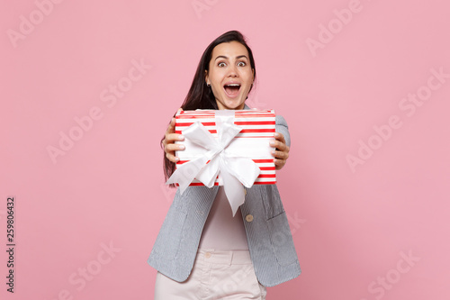 Surprised young woman holding red striped present box with gift ribbon isolated on pink pastel background. St. Valentine's Day International Women's Day, birthday, holiday concept. Mock up copy space.