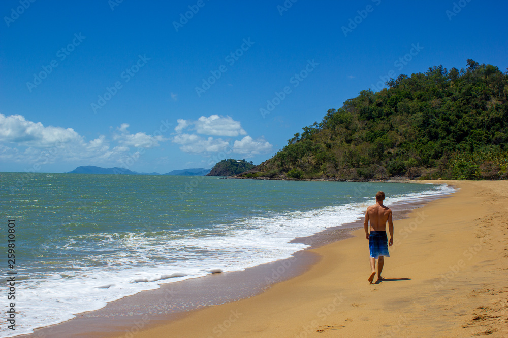 Muscular caucasian man walking on the beach as ocean waves crash behind him, cape tribulation, queensland, australia