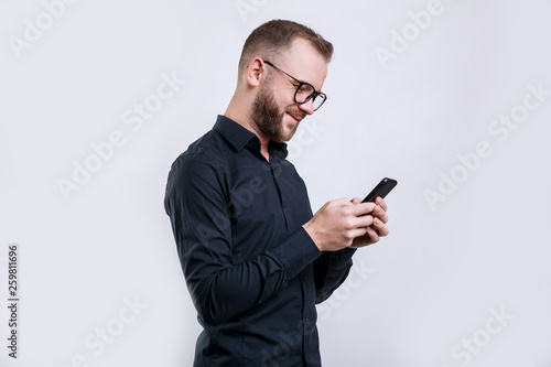 Portrait of young bearded caucasian man in glasses looking at mobile phone over white background,