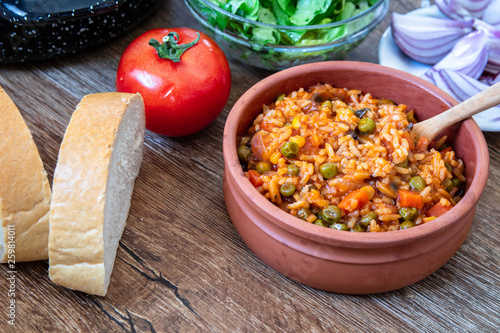 Fried rice with vegetables and spices in a vintage clay bowl and tomato, lettuce, onion and bread on wooden table