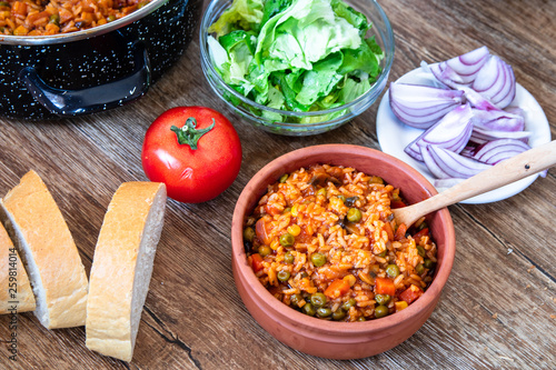 Fried rice with vegetables and spices in a vintage clay bowl and tomato, lettuce, onion and bread on wooden table