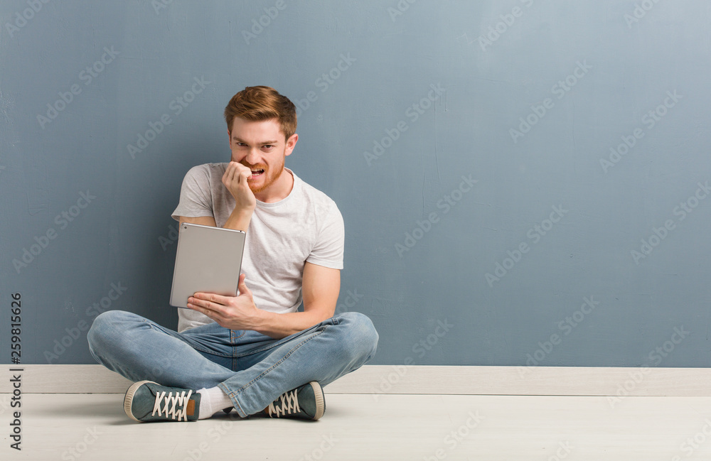Young redhead student man sitting on the floor biting nails, nervous and very anxious. He is holding a tablet. Young redhead student man sitting on the floor biting nails, nervous and very anxious. He is holding a tablet.
