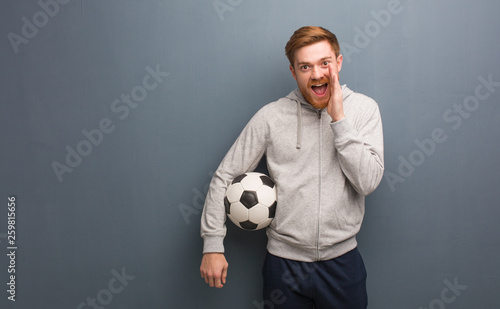 Young redhead fitness man shouting something happy to the front. He is holding a soccer ball.
