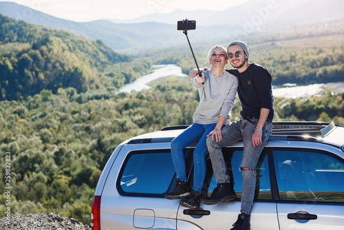 Young hipster couple in love making selfie using smart phone while sitting on the roof of off-road car on mountain background.