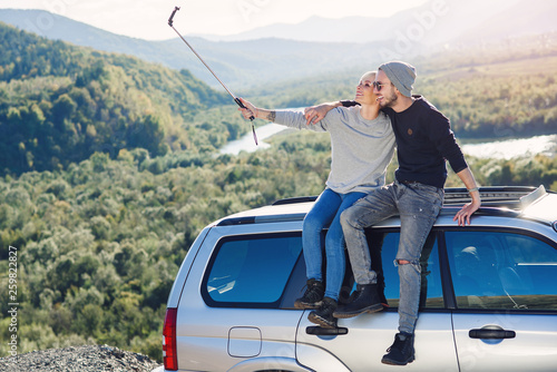 Young hipster couple in love making selfie using smart phone while sitting on the roof of off-road car on mountain background.