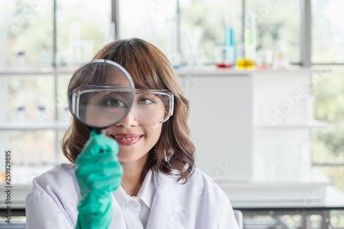 A young Asian woman scientist working in laboratory holding a magnifying glass to her face looking at camera.