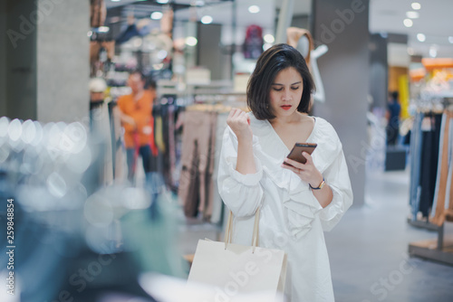 Young Asian beautiful woman shot hair shopping new clothes in a shopping mall store. Woman standing and holding a shopping bag and looking to her mobile phone in the store.