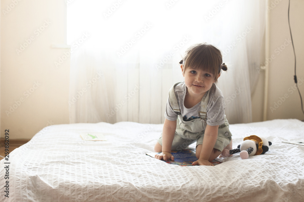 Cute toddler girl reading a book sitting in a white bedroom Cute toddler girl reading a book sitting in a white bedroom