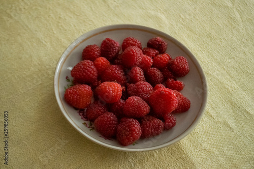 Ripe raspberries are laying on the white plate which is standing on the table covered with yellow tablecloth