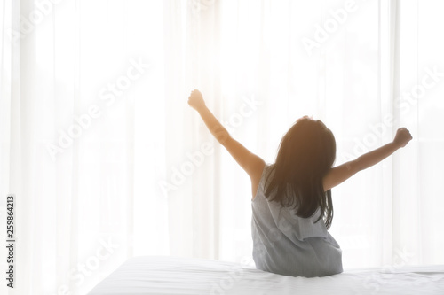 Asian child relax or kid girl wake up or woke up with stretch oneself after sleep for refreshing in morning on white bed and window with curtain in bedroom at home or hotel resort on holiday and warm