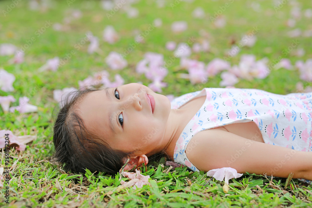 Smiling little girl lying on green grass with fall pink flower in the garden outdoor.