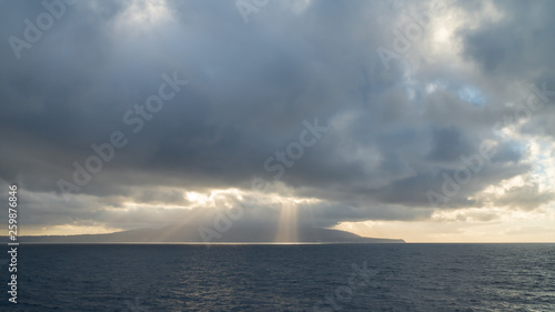 Sun rays burst out from behind clouds over the ocean © Keerathi