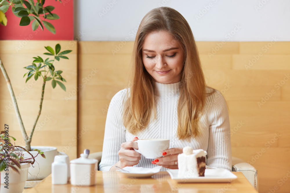 Attractive model sits in chair and looks in cup of coffee. Dreaming girl with blond hair wears white dress, holds cup in two hands, looks delighted, enjoys pleasant atmosphere of cosy cafe. Attractive model sits in chair and looks in cup of coffee. Dreaming girl with blond hair wears white dress, holds cup in two hands, looks delighted, enjoys pleasant atmosphere of cosy cafe.