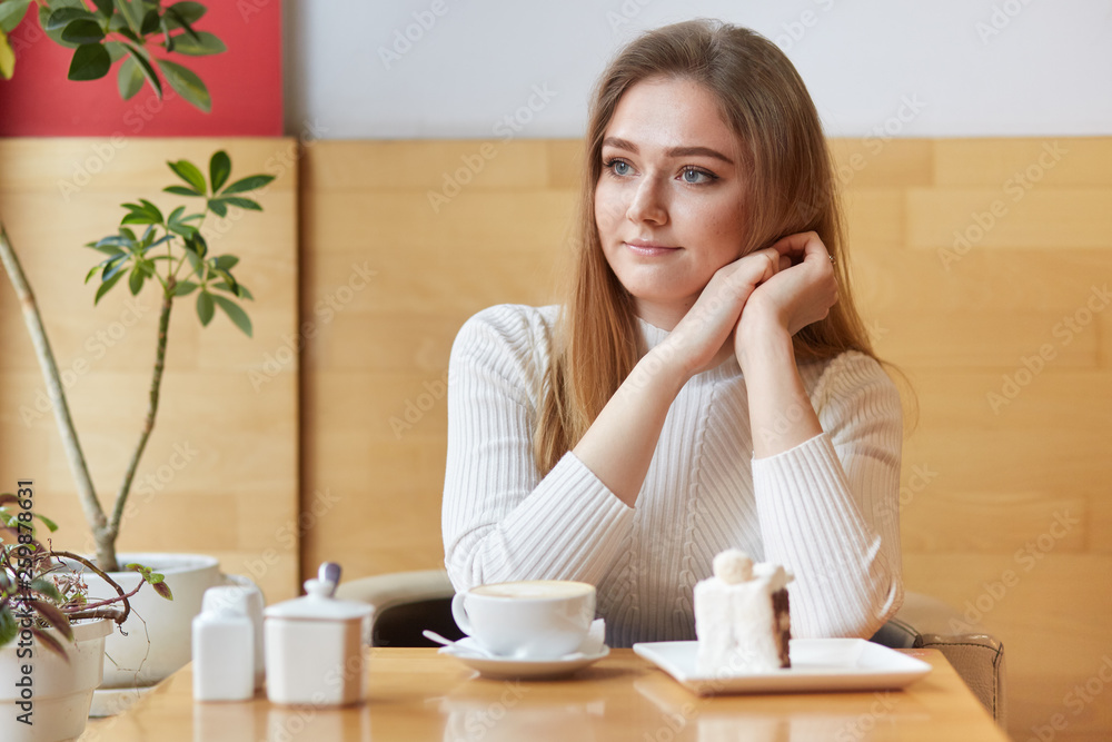 Beautiful model sits in local cafe, looks in window, eats delicious cake and drinks coffee, puts her head on hands, feels satisfied. Tender young girl having rest in cosy and pleasant atmosphere. Beautiful model sits in local cafe, looks in window, eats delicious cake and drinks coffee, puts her head on hands, feels satisfied. Tender young girl having rest in cosy and pleasant atmosphere.