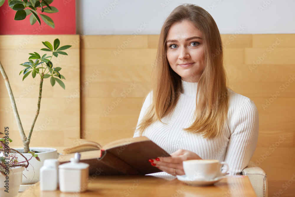 Adorable blond visitor of local cafe wears white dress, eats creamy tasty cake, drinks cup of coffee. Sincere cute model poses with brown old book in her hands. Free time activities concept. Adorable blond visitor of local cafe wears white dress, eats creamy tasty cake, drinks cup of coffee. Sincere cute model poses with brown old book in her hands. Free time activities concept.