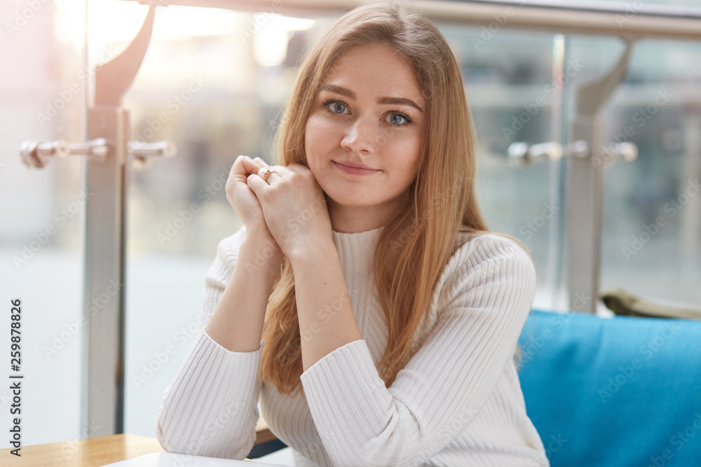Portrait of attractive young girl being in good mood, has rest in cafeteria after lectures in university, sits on comfortable blue sofa, wears casually, looks at camera, keeps hands folded under chin. Portrait of attractive young girl being in good mood, has rest in cafeteria after lectures in university, sits on comfortable blue sofa, wears casually, looks at camera, keeps hands folded under chin.