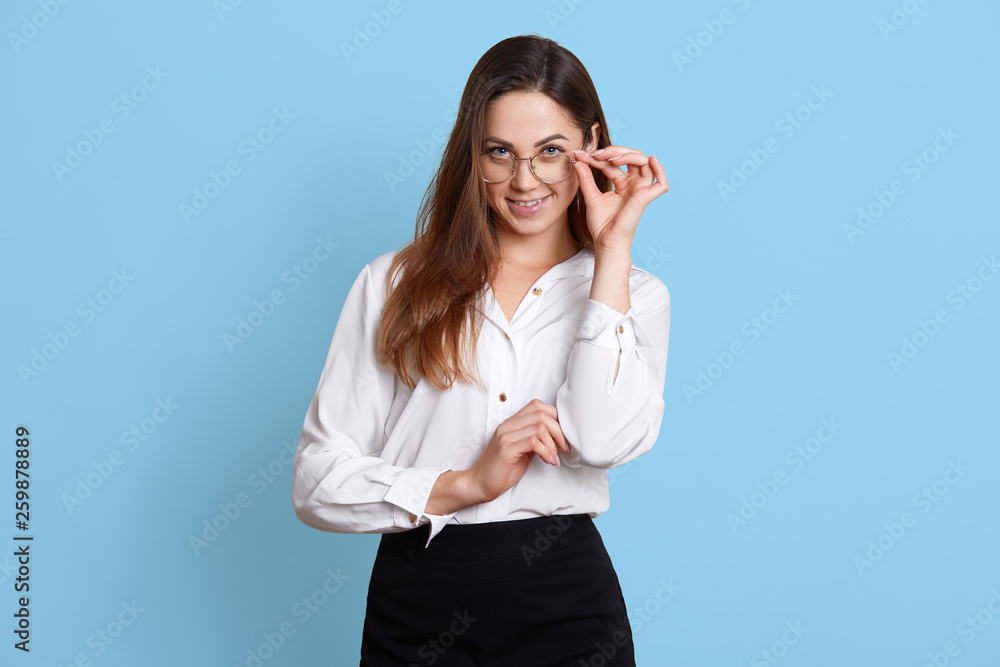 Portrait of standing cheerful businesswoman in glasses, touching them with her fingers, isolated over light blue background. Smiling long haired woman wears office uniform. Office work concept. Portrait of standing cheerful businesswoman in glasses, touching them with her fingers, isolated over light blue background. Smiling long haired woman wears office uniform. Office work concept.