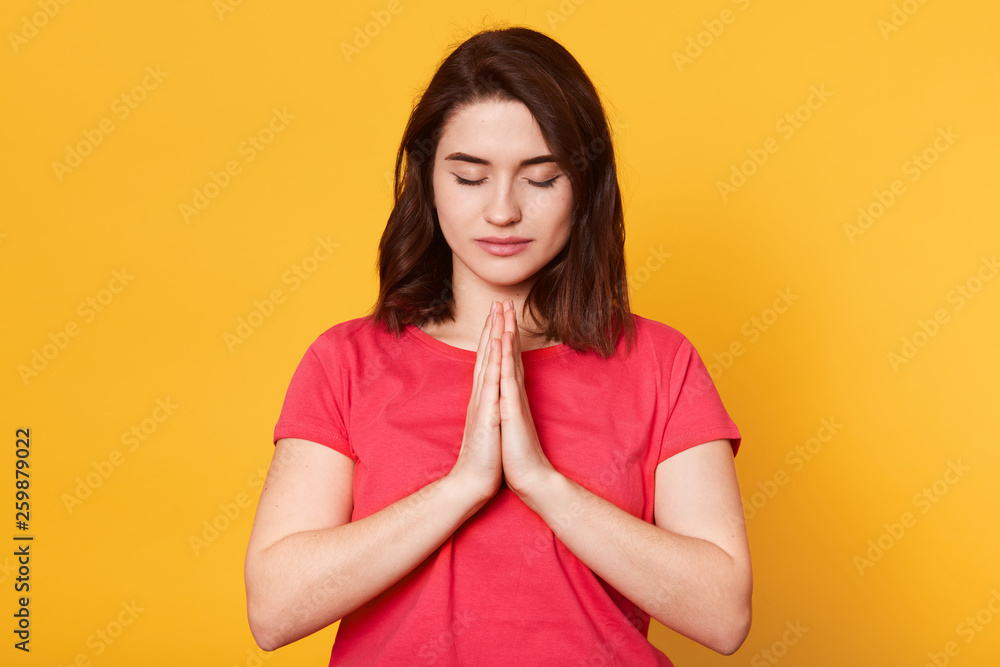 Beautiful young brunette female makes praying gesture, keeps palms pressed together, feels guilty, dressed in casual redt shirt, asks for apologize, isolated over yellow background. People concept. Beautiful young brunette female makes praying gesture, keeps palms pressed together, feels guilty, dressed in casual redt shirt, asks for apologize, isolated over yellow background. People concept.
