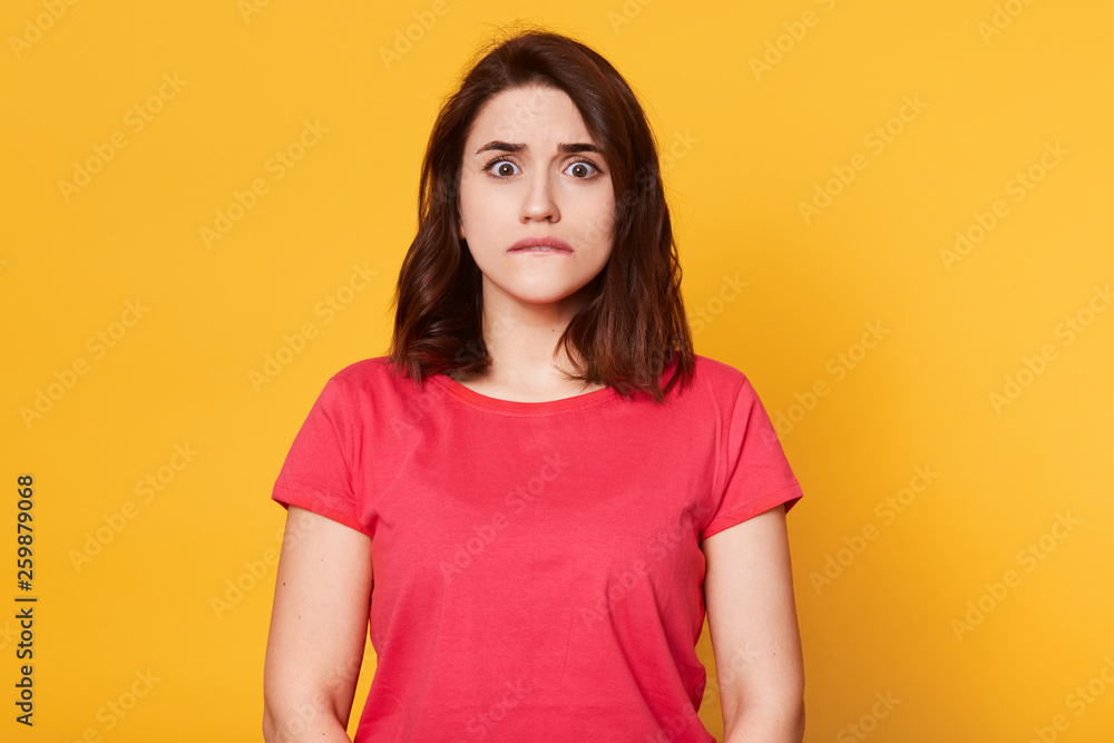 Portrait of young pretty emotional woman on yellow background, bites her lip, looks at camera with scared facial expression, afraids of something, warches horror film. People and emotions concept. Portrait of young pretty emotional woman on yellow background, bites her lip, looks at camera with scared facial expression, afraids of something, warches horror film. People and emotions concept.