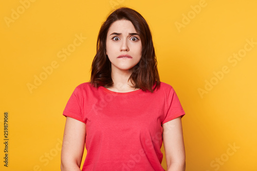 Portrait of young pretty emotional woman on yellow background, bites her lip, looks at camera with scared facial expression, afraids of something, warches horror film. People and emotions concept.