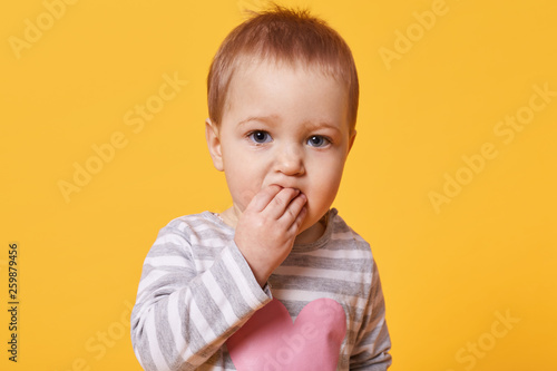 Portrait of a cute brooding girl with short fair hair keeping fingers in her mouth. A serious kid stands in front of camera looking straight  feels bored  search for new funny pastime. Family concept.