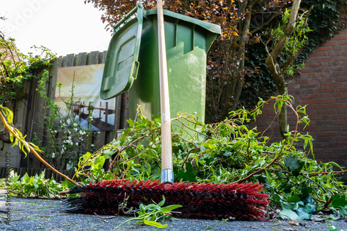 Green wheelie bin / garden waste container and broom filled with fruit and vegetable waste, garden waste, organic waste for composting and fermentation. Recycling garbage for a better environment.