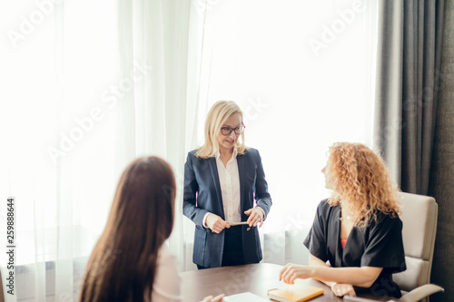 Female colleagues discussing work plans together, sitting at office table. Female red-haired intern asks questions to blonde head of department. Chief Executive explaining employees working tasks.