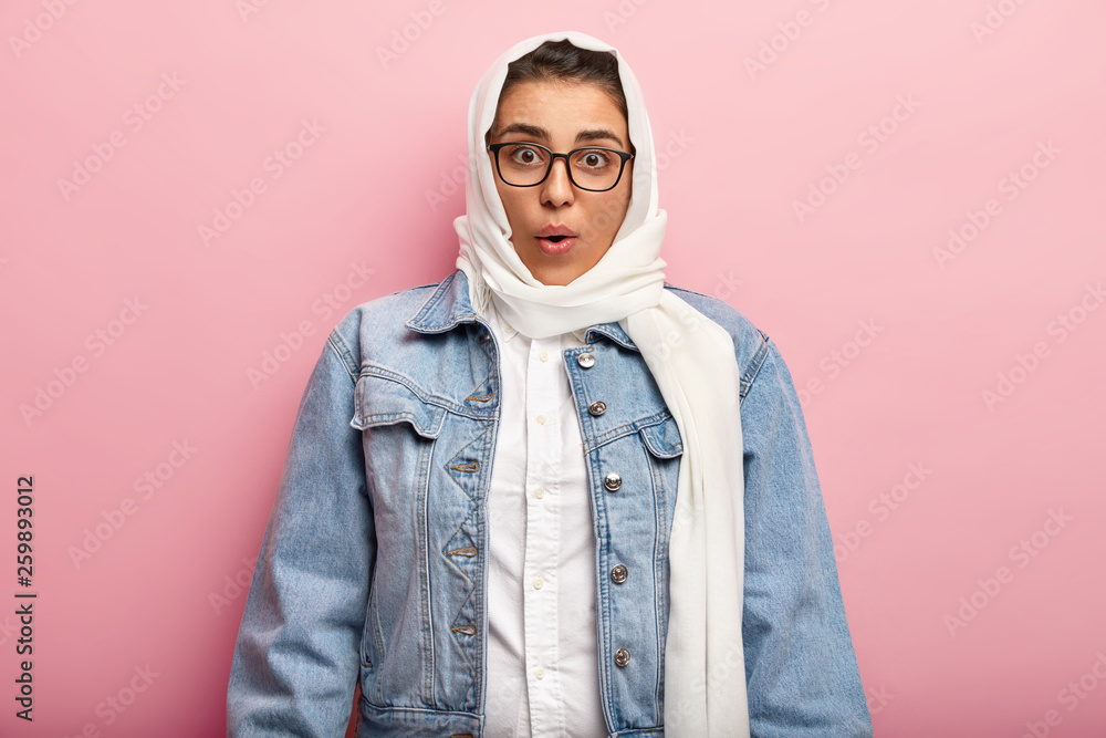 Suprised Muslim woman covers head with hijab, wears square glasses and denim jacket, being very religious, shocked by strict rules of religion, isolated over pink background. Ethnicity concept Suprised Muslim woman covers head with hijab, wears square glasses and denim jacket, being very religious, shocked by strict rules of religion, isolated over pink background. Ethnicity concept