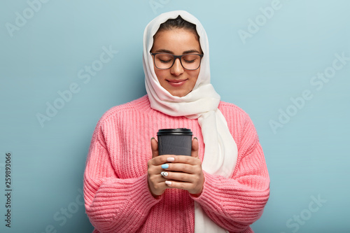 Pleased Arabic lovely woman warms hands on hot paper cup with drink, enjoys morning coffee, has gentle friendly expression, wears white scarf on head, loose pink jumper, poses indoor. Spare time © wayhome.studio 