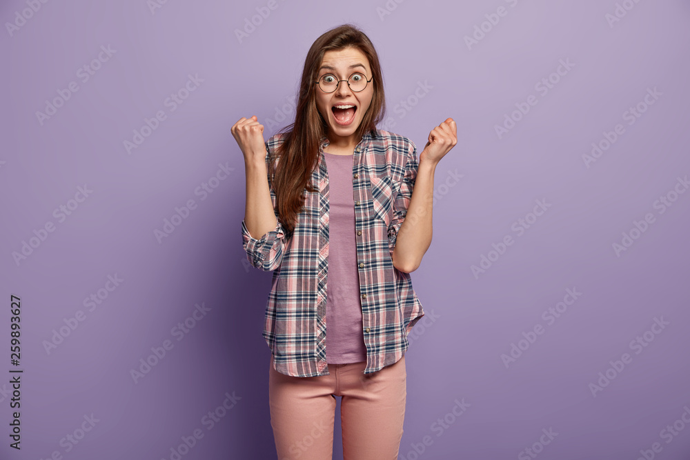 Brunette happy girl clenches fists like winner, has surprised overjoyed facial expression, keeps mouth opened, wears stylish checkered shirt, poses and gestures over purple background, got victory Brunette happy girl clenches fists like winner, has surprised overjoyed facial expression, keeps mouth opened, wears stylish checkered shirt, poses and gestures over purple background, got victory