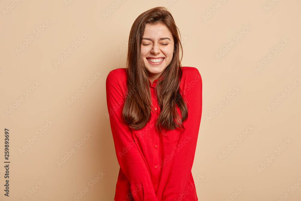 Isolated shot of positive young European woman feels enjoyment and pleasure, keeps eyes closed, smiles broadly, dressed in red clothes, isolated over brown background. People, emotions concept Isolated shot of positive young European woman feels enjoyment and pleasure, keeps eyes closed, smiles broadly, dressed in red clothes, isolated over brown background. People, emotions concept