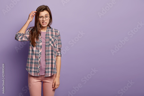 Photos Horizontal shot of confused European young woman scratches head, has indecisive