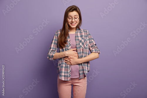 Canvas Print Glad woman has full stomach after delicious dinner, keeps both hands on belly, being well fed smiles positively dressed in casual clothes round spectacles isolated over purple wall