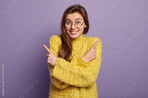 Glad lovely dark haired woman points fingers to both sides, shows different direction, wears yellow sweater, chooses between two objects, isolated over purple background. Look at empty corners photo