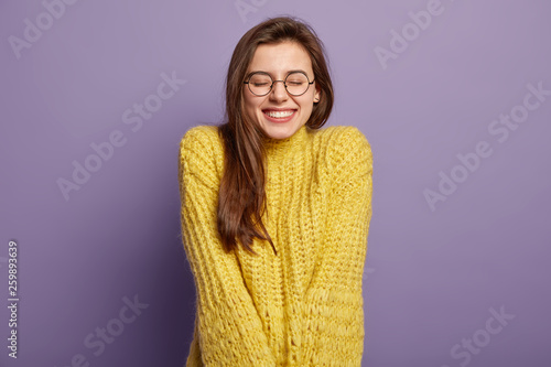 Isolated portrait of happy woman has toothy smile, closes eyes, feels pleasure from good compliment, wears glasses and yellow jumper, stands over purple wall. Positive emotions and feelings concept