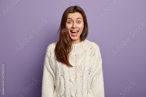 Positive young female with glad expression, opens mouth from surprise, reacts on positive unexpected news, wears white jumper, stands against violet studio wall, intrigued by telling something