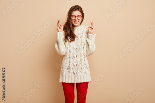 Indoor shot of glad woman with glad facial expression, keeps fingers crossed, hopes dreams come true, wears long sweater and pantyhose, isolated against beige background. Believe only in better!