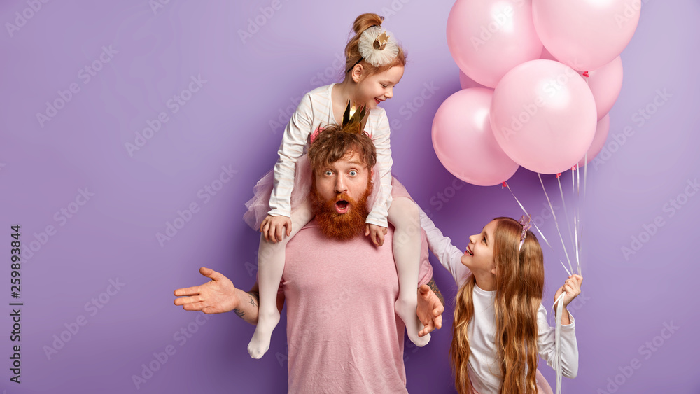 Fototapeta premium Indignant foxy father left alone with small children, plays with daughters, organizes real party for children, has uncertain expression. Two female kids play together, hold pink air balloons